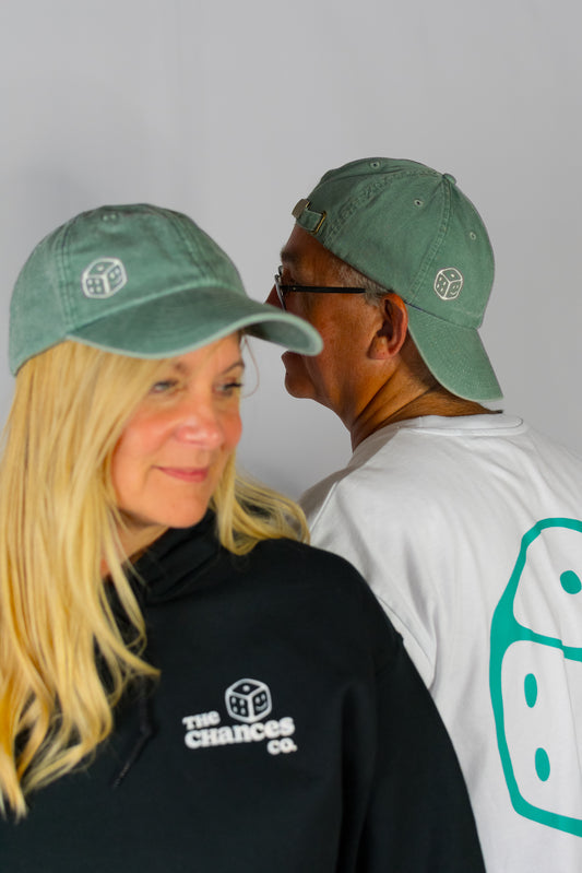 Two people wearing green caps with a smiley dice logo on and black shirts with 'The Chances Co.' logo on a grey background