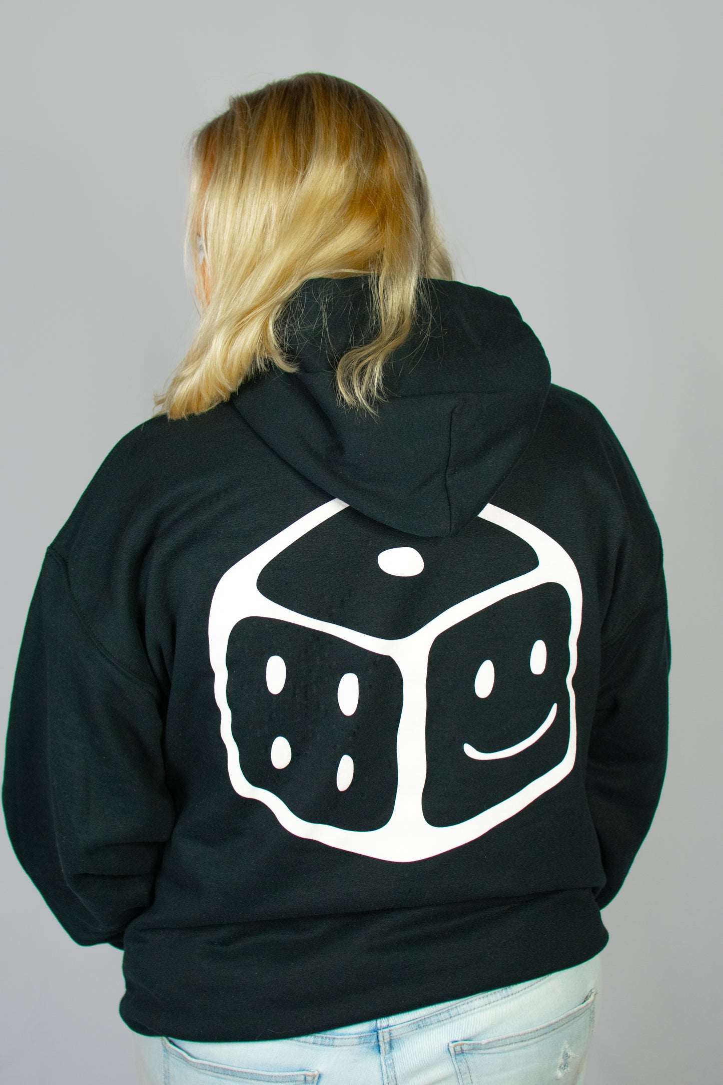 Person wearing a black hoodie with a white smiley dice design on the back against a grey background