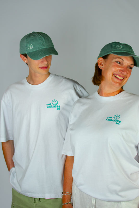 Two people smiling while wearing green caps and white t-shirts with the Chances Clothing smiley dice logo on the front. In front of a plain background