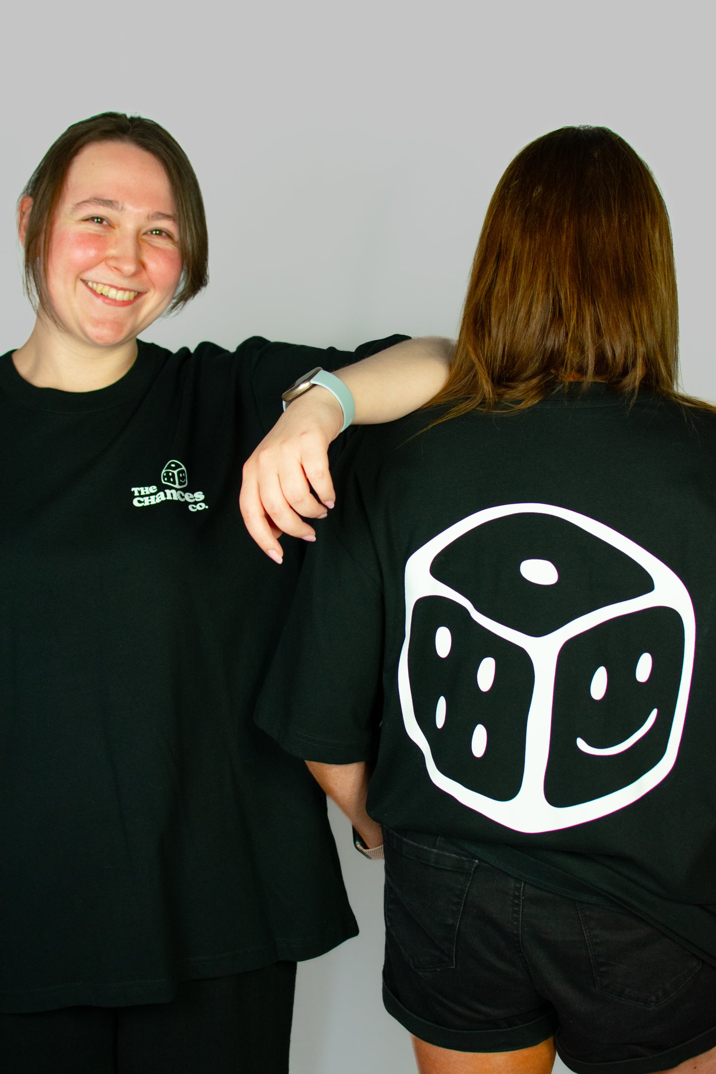 Two people wearing black t-shirts, one with the white Chances dice design on the back. One with a small white smiley dice and the chances co written on the front
