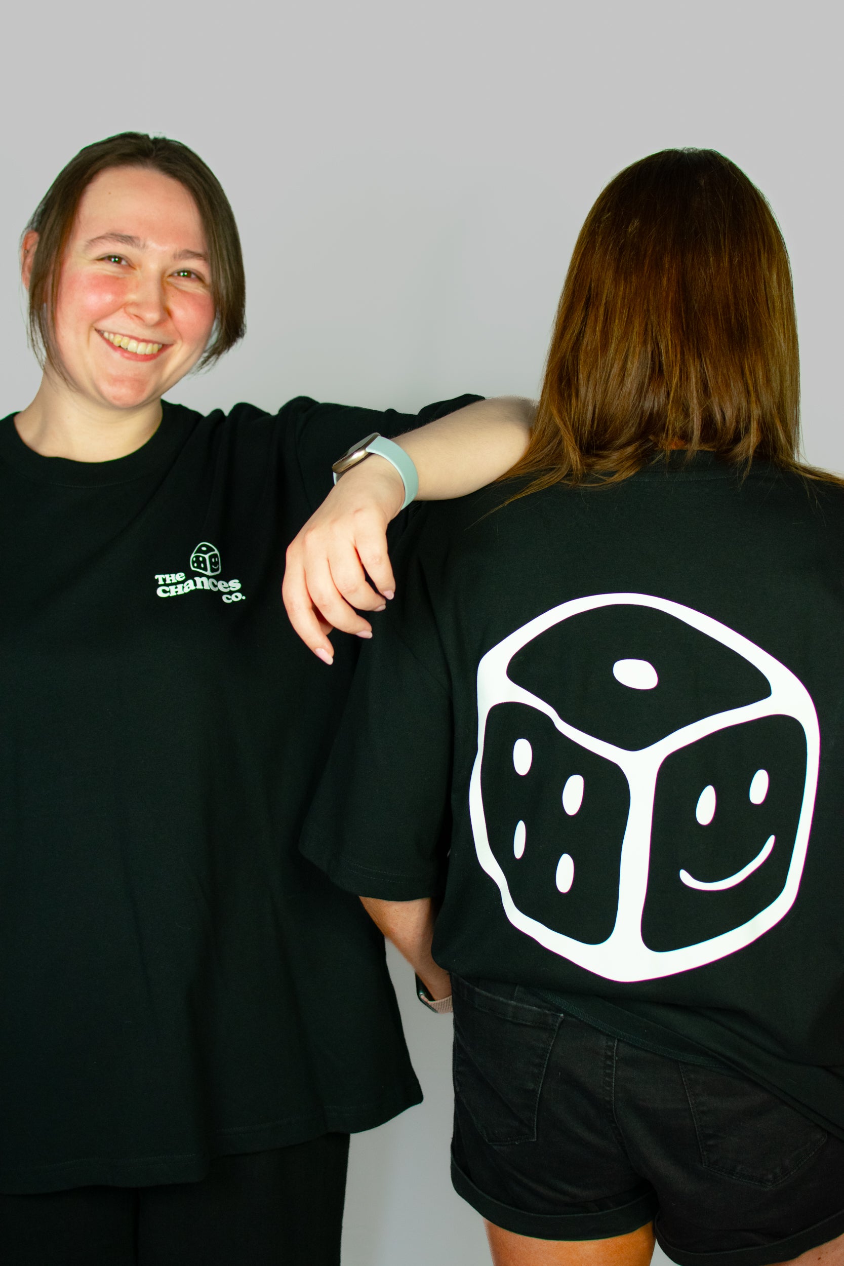Two people wearing black t-shirts, one with the white Chances dice design on the back. One with a small white smiley dice and the chances co written on the front