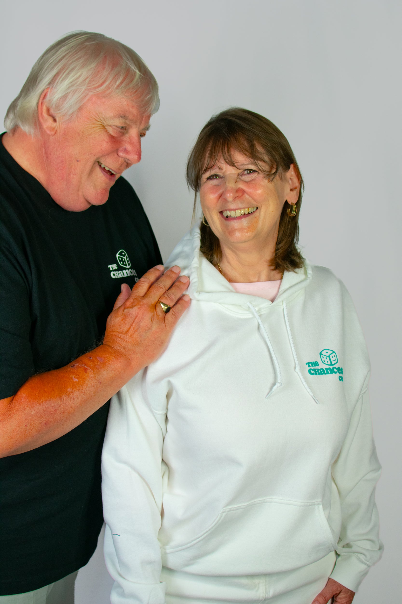 Two elderly people wearing a white hoodie and a black tee with a smiley dice logo stood in front of a grey background