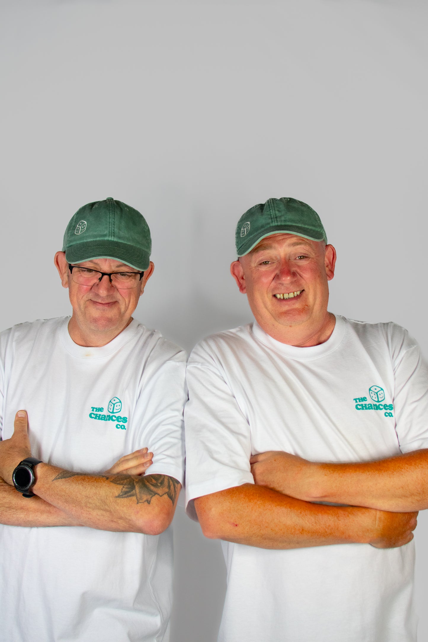 Two men wearing green caps and white shirts with a green smiley dice logo on both the caps and T-shirts . Stood in front of a plain background