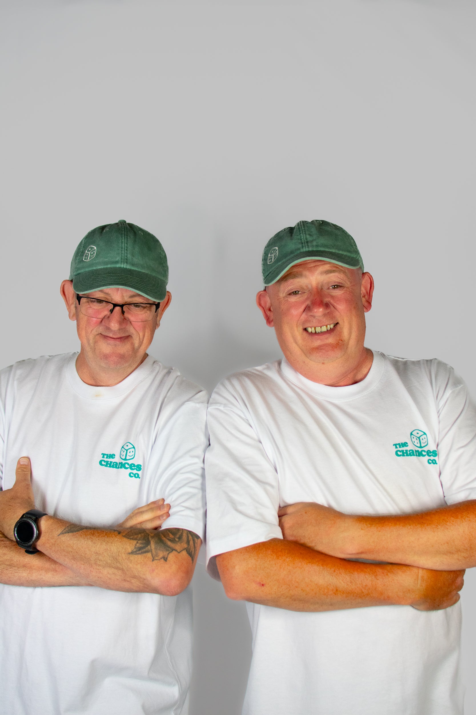Two men wearing green caps and white shirts with a green smiley dice logo on both the caps and T-shirts . Stood in front of a plain background