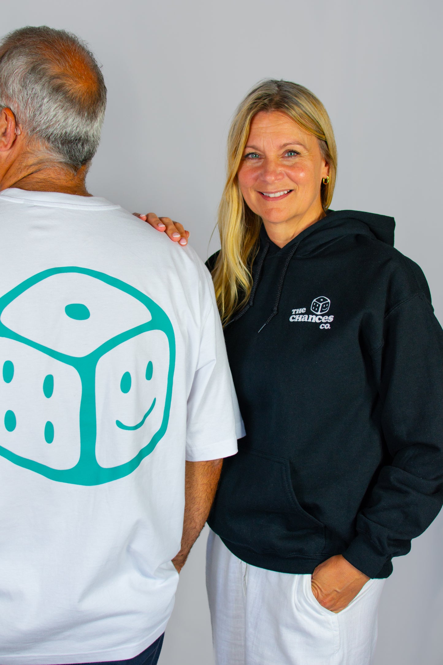 Person wearing a white shirt with a green smiley dice design, standing next to another person in a black hoodie with a white smiley dice and "The Chances Co" logo.