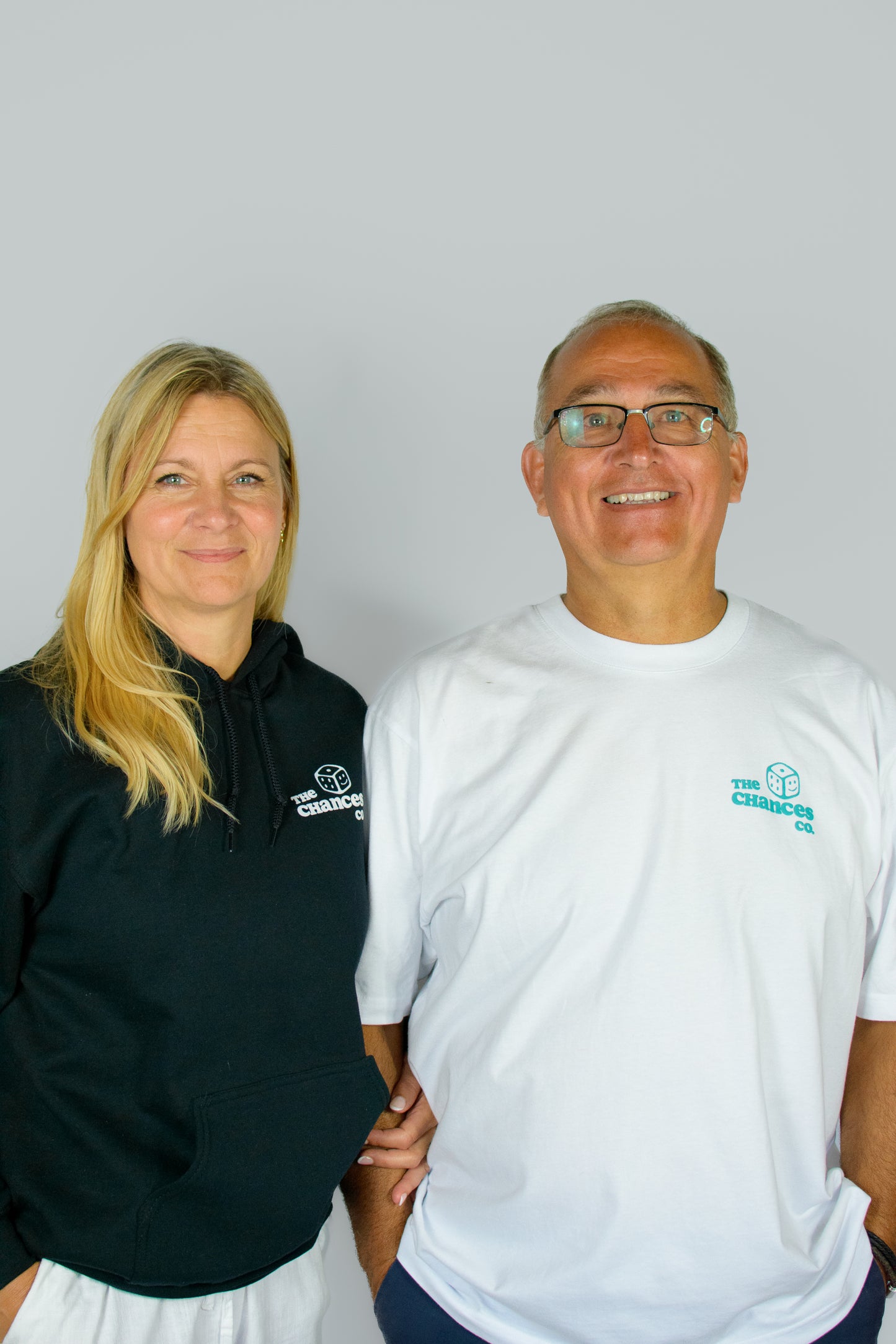 Two people stood wearing a white Chances Clothing graphic tee with the green dice logo and one in a black hoodie with the white smiley face logo.
