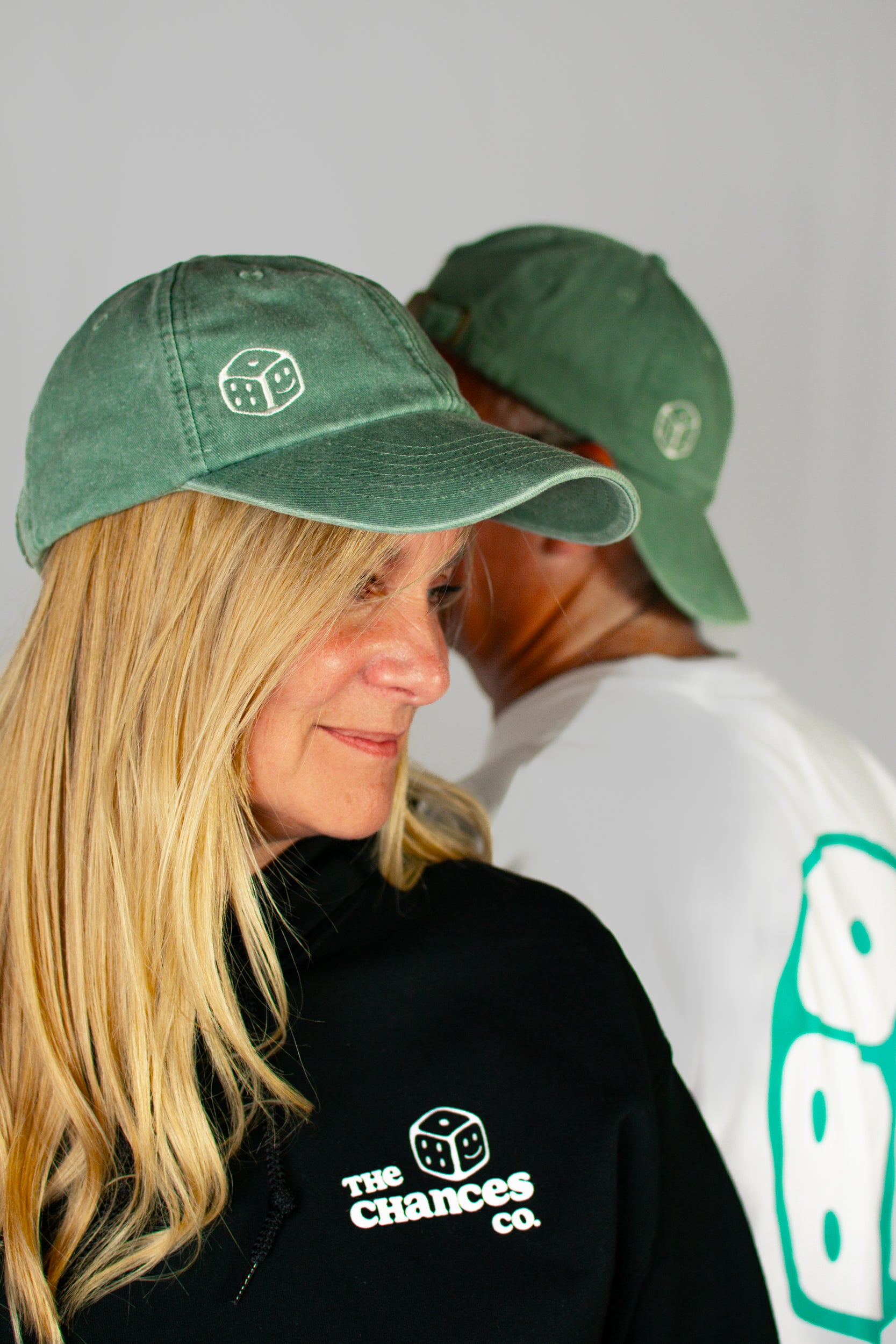 Two people wearing green caps with a smiley dice logo on and black shirts with 'The Chances Co.' logo on a grey background