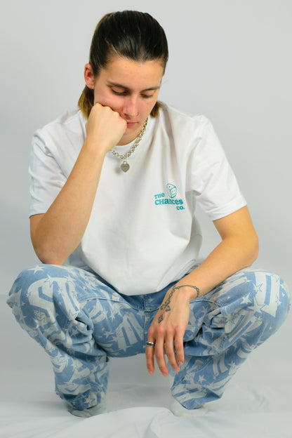 Person wearing a white t-shirt with a green smiley dice logo and blue patterned pants on a plain background