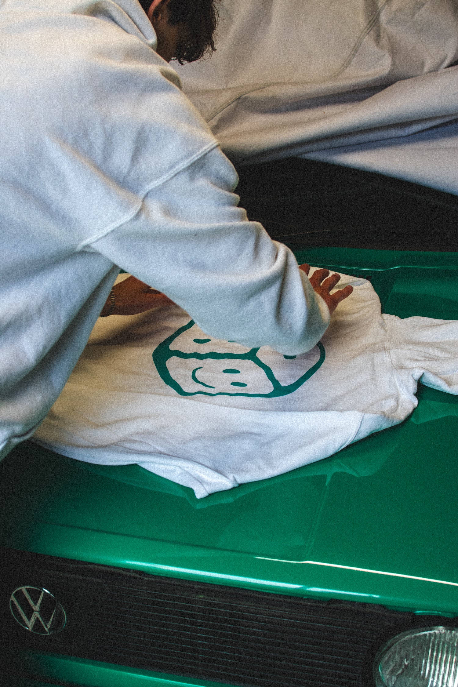 Person folding a Chances Clothing Hoodie on the bonnet of a car. It has a green smiley dice logo on the back of it. 