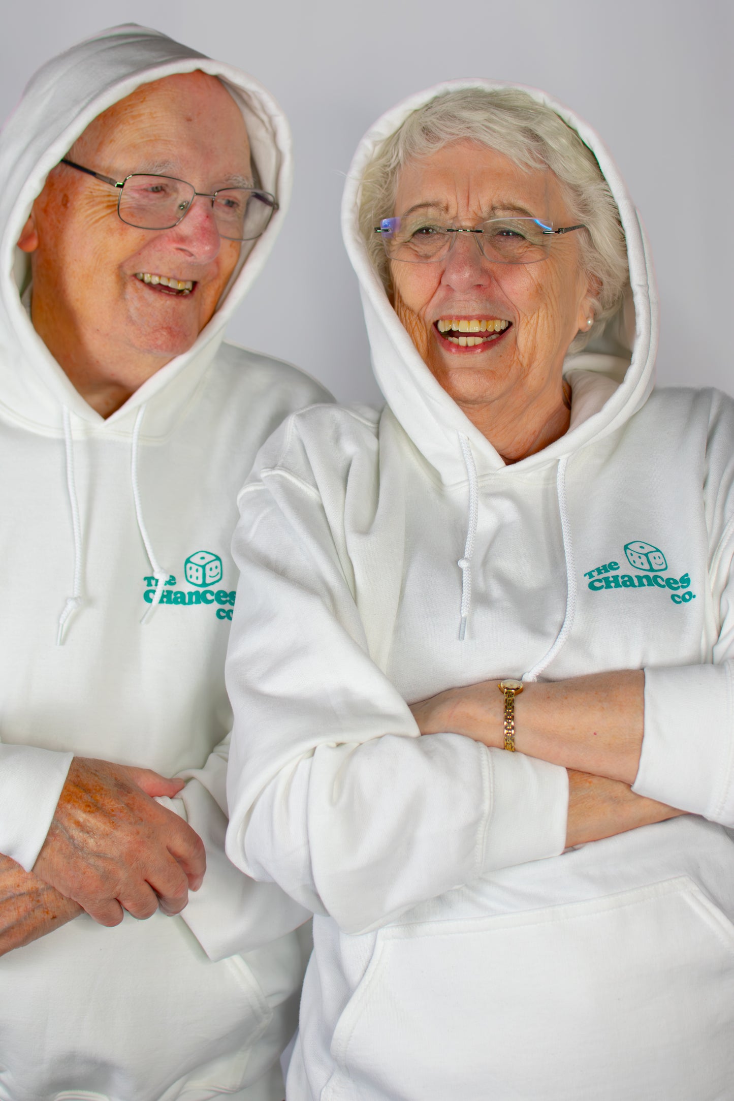 Two elderly people wearing matching Chances Clothing hoodies with a green smiley dice logo with "the chances co" written next to it.