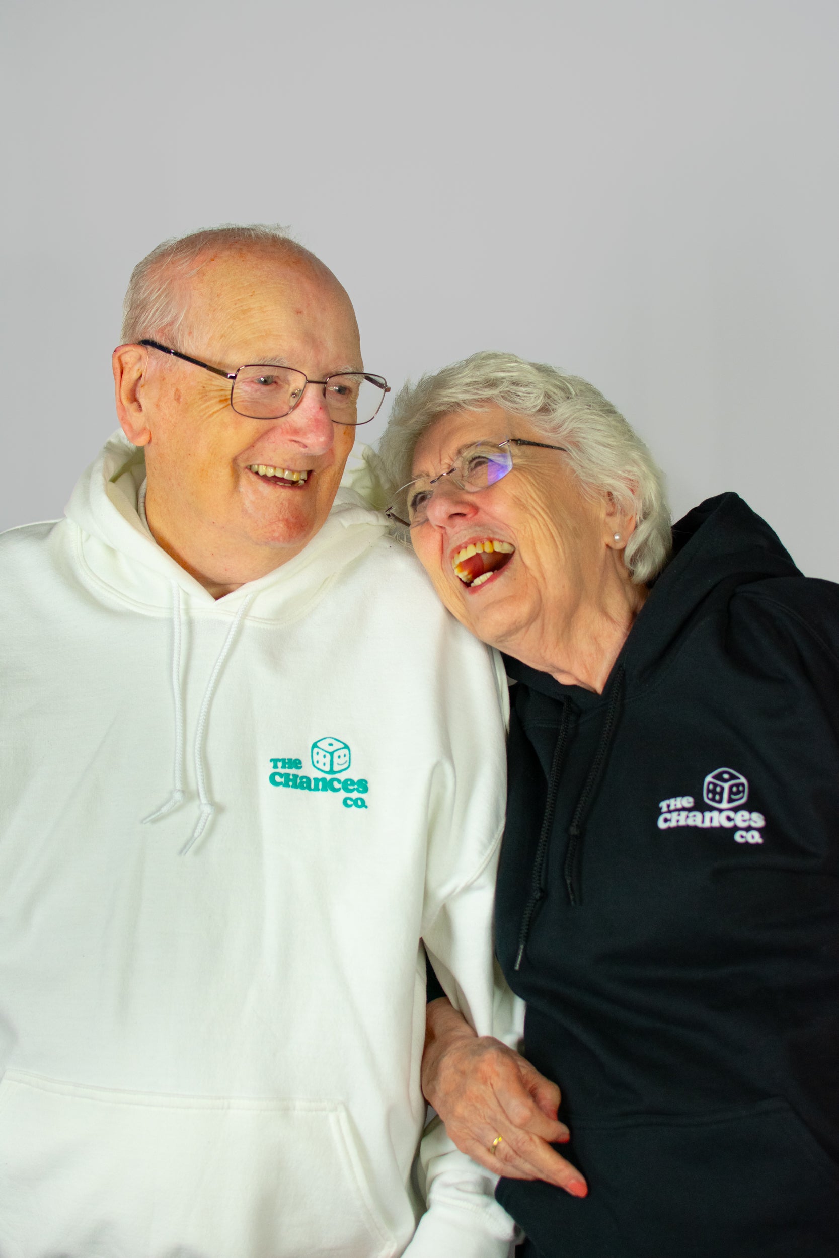 Two elderly people wearing white and black hoodies with a small smiley dice next to "the Chances co" logo on a plain background