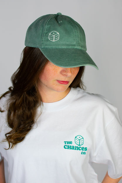 Person wearing a green cap with a smiley dice logo on and a white t-shirt with the same Chances Clothing smiley dice logo on a grey background looking down