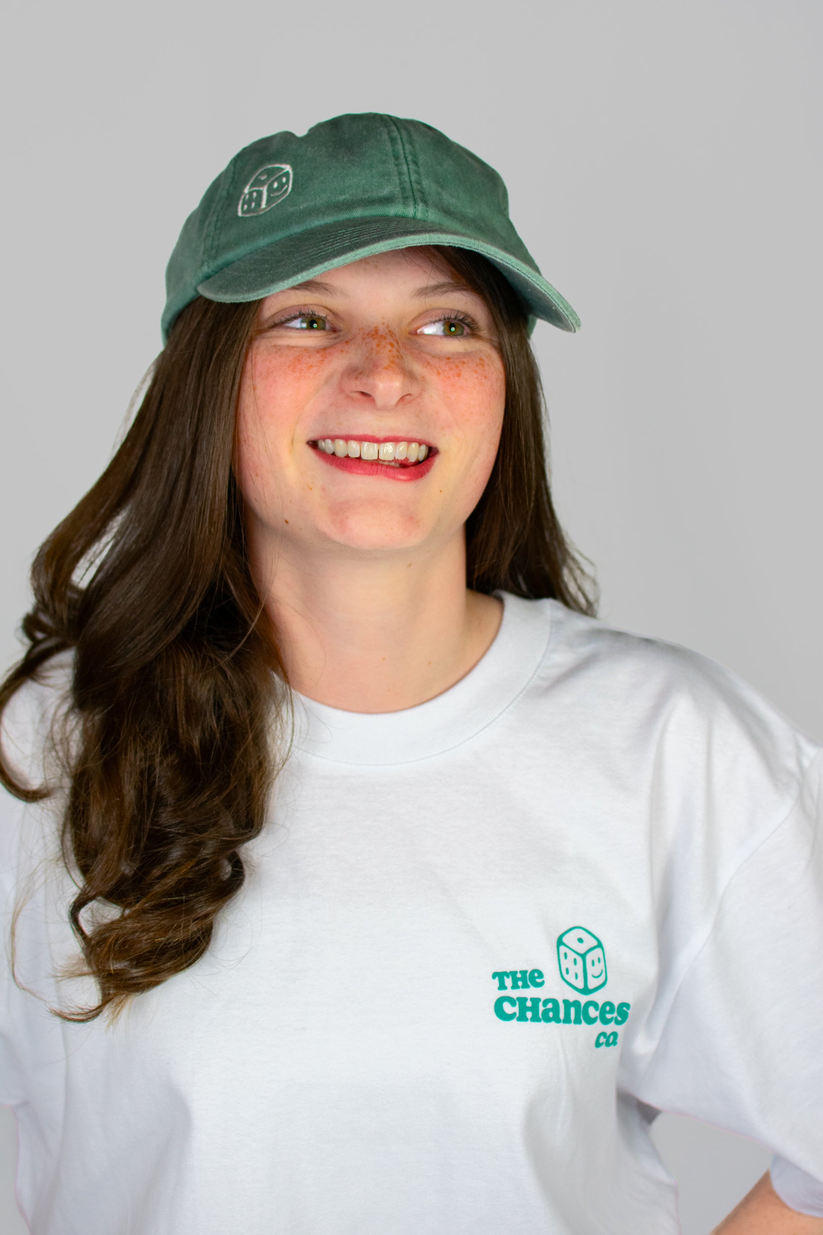 Person wearing a green cap with a smiley dice logo on and a white t-shirt with the same Chances Clothing smiley dice logo on a grey background looking to the left
