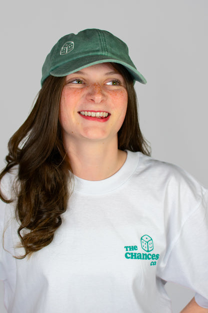 Person wearing a green cap with a smiley dice logo on and a white t-shirt with the same Chances Clothing smiley dice logo on a grey background looking to the left