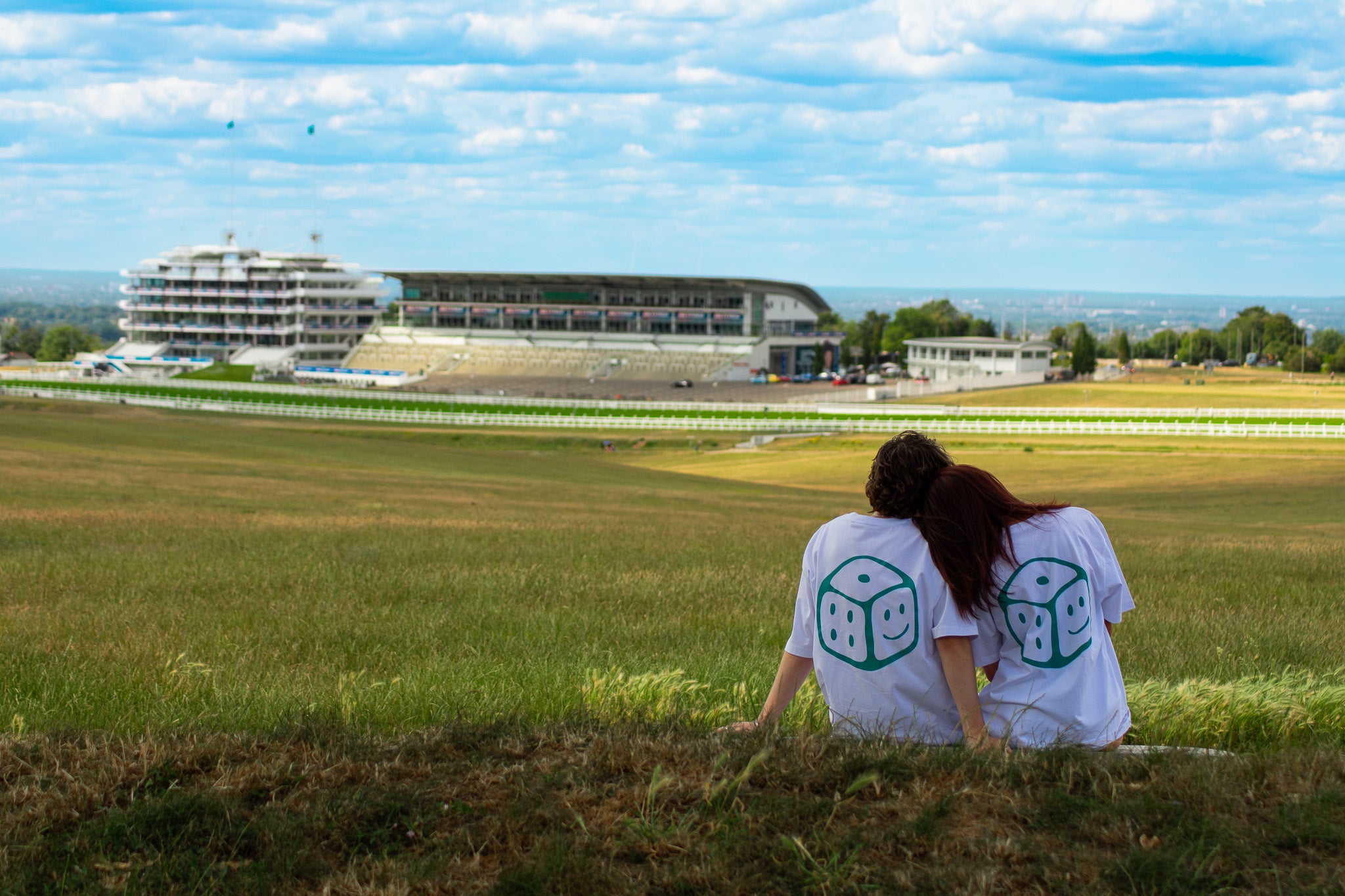 Two People sat on grass wearing chances clothing t-shirts with a green smiley dice on the back of the t-shirt