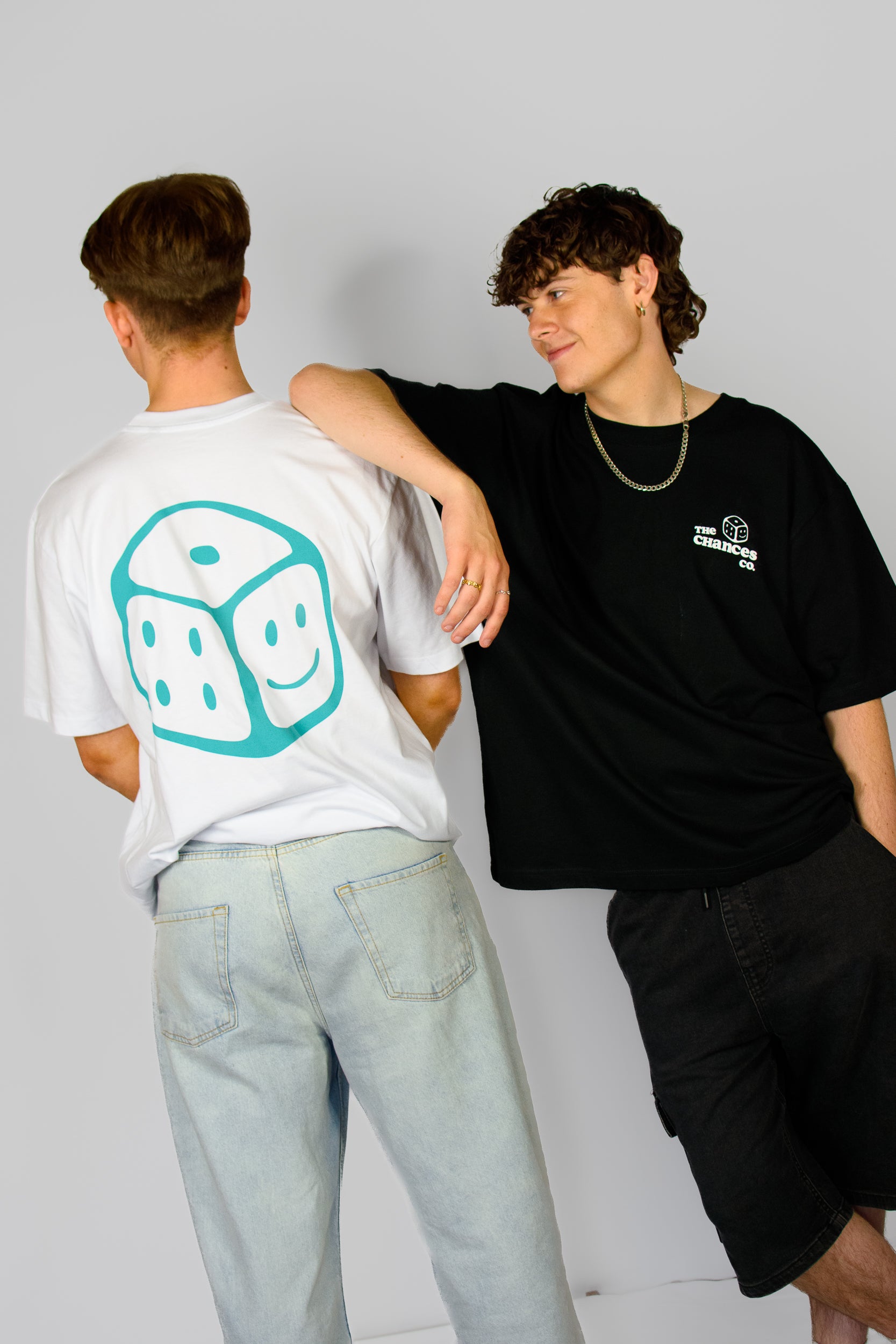 Two people wearing t-shirts with Chances Clothing logos, one white and one black, on a plain background. the white tee has a green smiley dice on the back