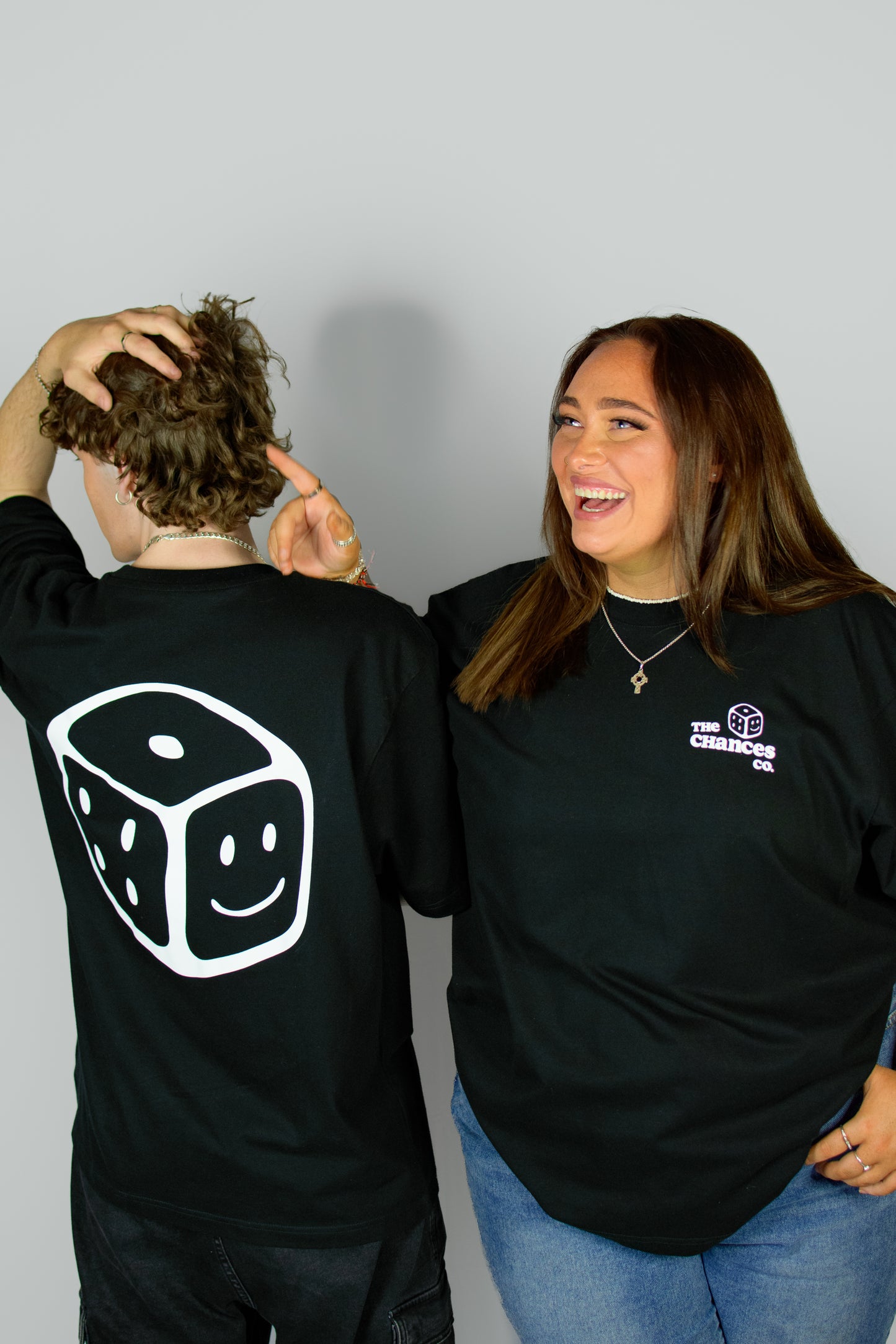 Two people wearing black t-shirts with logos, one featuring a white smiley dice design on the back. One with "the chances co" and a dice on the front