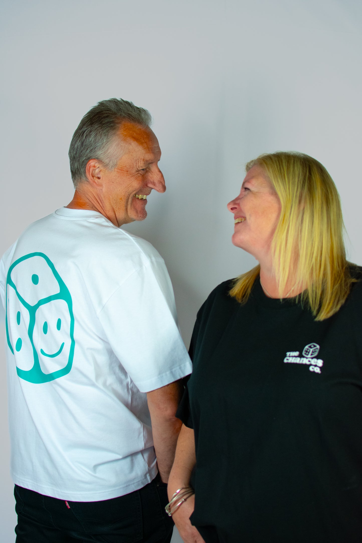 Two people standing close together against a plain background. One is wearing a white T-shirt with a green smiley face dice on the back and one is wearing a black tee with "The Chances Co" on the front