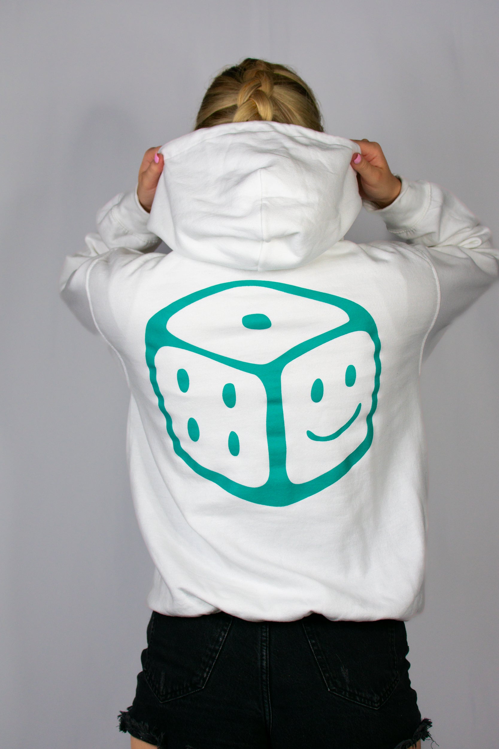Person wearing a white hoodie with a teal smiley dice design on the back against a grey background