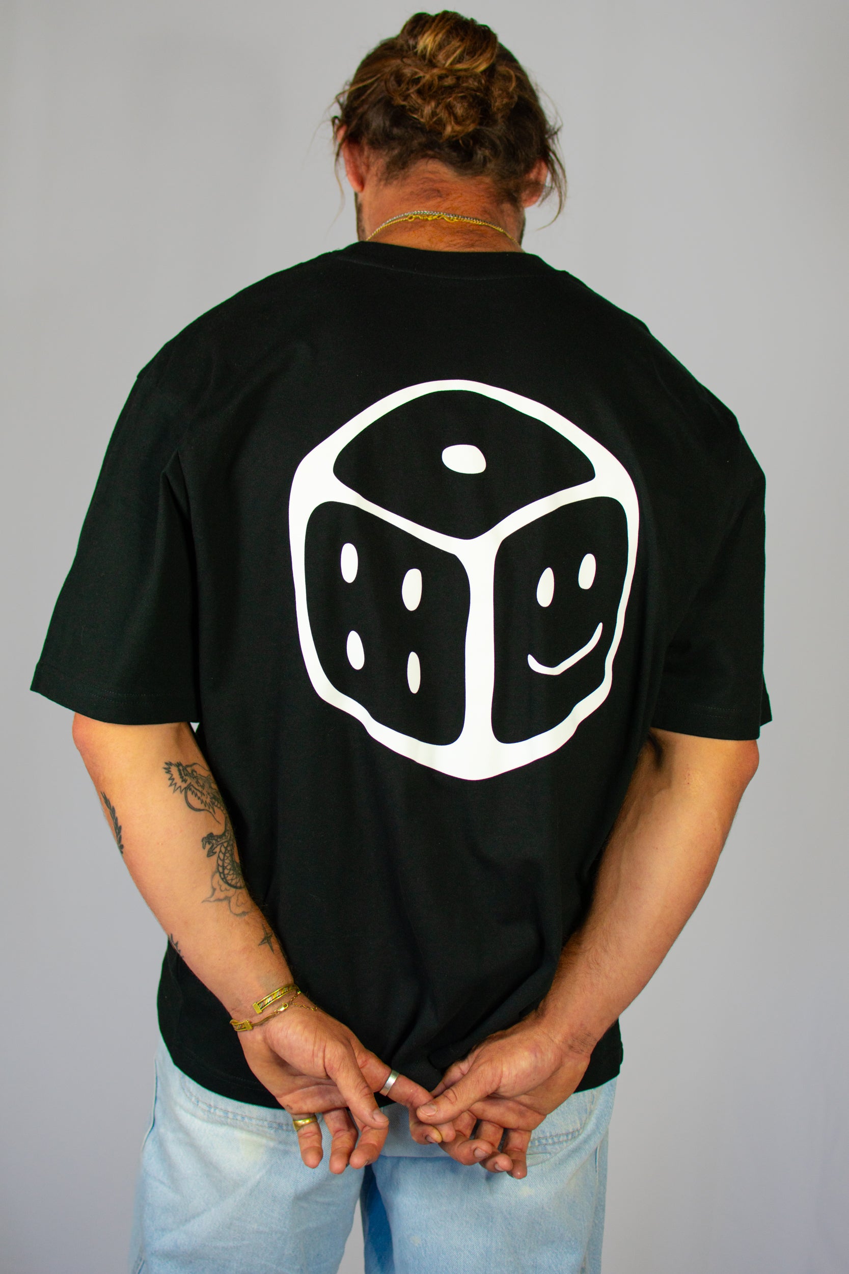 Person wearing a black t-shirt with a white smiley dice design on the back, standing against a plain background.