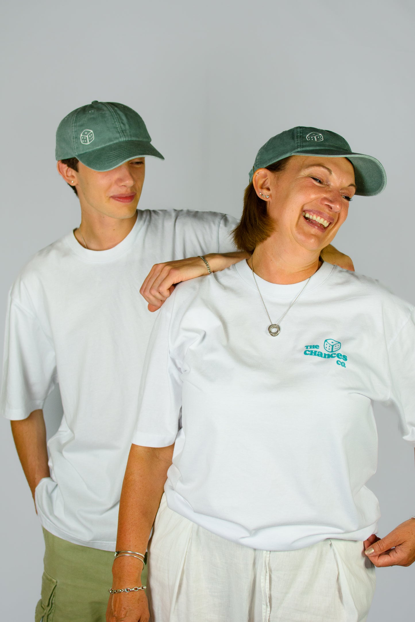 Two people wearing green caps and white t-shirts with the teal smiley dice chances clothing logo, standing close together and laughing on a plain background.