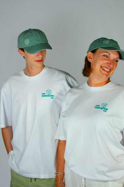 Two people smiling while wearing green caps and white t-shirts with the Chances Clothing smiley dice logo on the front. In front of a plain background