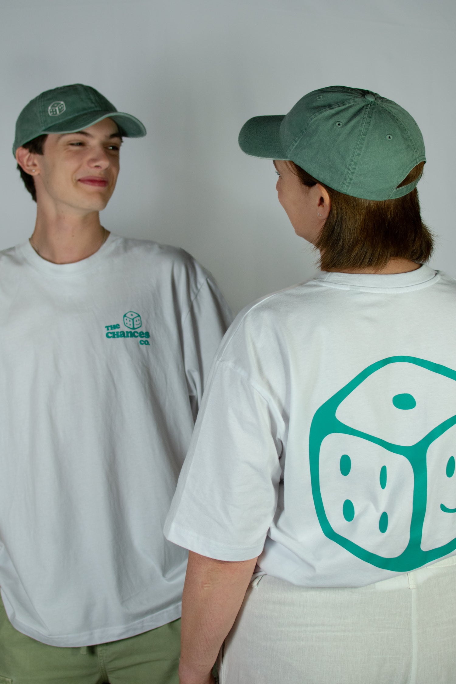 Two people wearing green caps and white t-shirts with Chances Clothing smiley dice logos, one facing forward and one facing backward. 