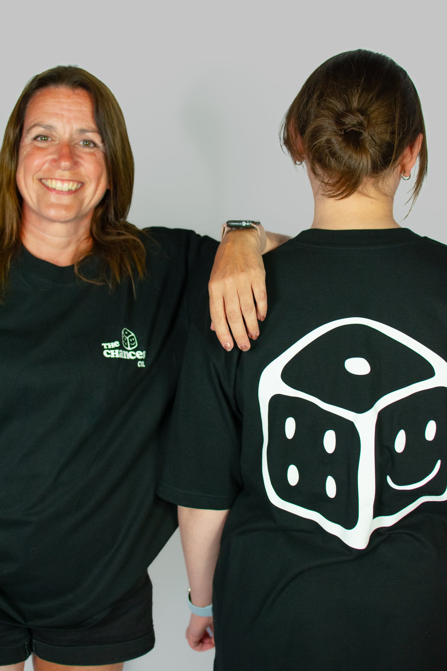 Two people wearing black t-shirts, one with a white smiley dice design on the back. One with "the Chances Co2 logo on the front and a small dice